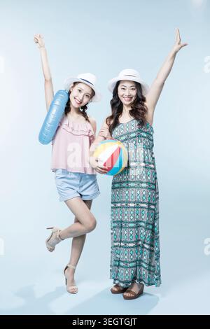 Zwei junge Frauen in Sommerhüten, eine mit erhobenen Armen und die andere mit einem Strandball, die zusammen auf hellblauem Hintergrund steht. Stockfoto