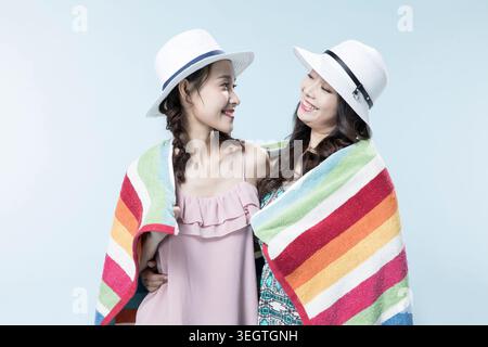 Zwei junge Frauen, lächelnd und in ein buntes Strandtuch gewickelt, stehen zusammen an einem sonnigen Strand. Stockfoto