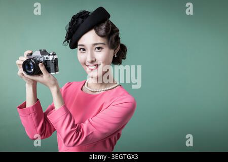 Eine junge Frau in einem rosafarbenen Kleid lächelt, während sie eine Kamera an ihr Auge hält und in einem modernen Studio für ein Foto posiert. Stockfoto