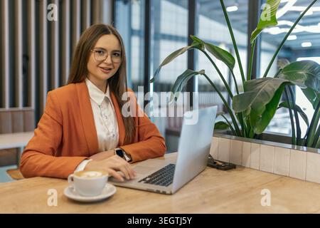 Selbstbewusste Geschäftsfrau, die im Café am Laptop arbeitet, Brillen trägt, Kaffee auf dem Tisch, modernen Arbeitsplatz, professioneller Lebensstil Stockfoto