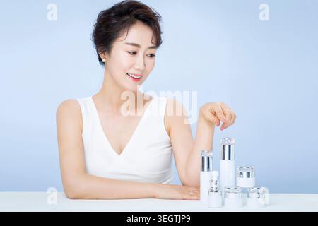 Eine Frau sitzt an einem Tisch, umgeben von verschiedenen Hautpflegeprodukten und lächelt, während sie sich darauf vorbereitet, eine Feuchtigkeitscreme aufzutragen. Stockfoto