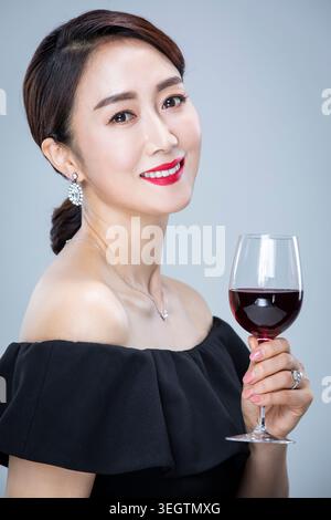 Eine Frau in einem schwarzen Kleid hält ein Glas Rotwein und lächelt in die Kamera. Stockfoto