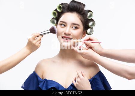 Einer Frau wird durch mehrere Hände geholfen, wobei eine Hand einen Pinsel hält, eine einen Make-up-Kompakt hält und andere Make-up-Produkte um ihr Gesicht halten. Stockfoto