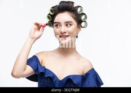 Eine junge Frau trägt Eyeliner auf ihr linkes Auge, während sie im Studio Haarrollen trägt. Stockfoto