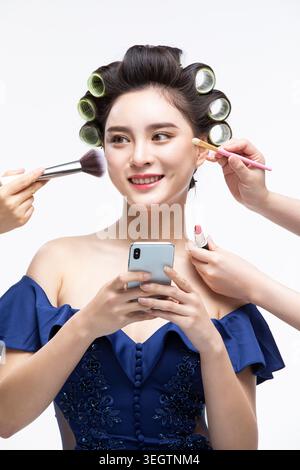 Eine Frau ist von mehreren Händen umgeben, die Kosmetikwerkzeuge und -Produkte halten, während sie ein Telefon hält und lächelt. Stockfoto