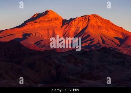 Blick aus der Vogelperspektive auf majestätische Berggipfel, die im warmen Licht des Sonnenuntergangs über der trockenen Wüstenlandschaft Toconao, Antofagasta, Chile leuchten. Stockfoto