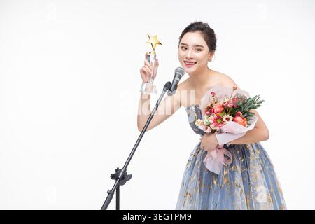 Eine junge Frau in einem formellen Kleid hält eine Trophäe und lächelt, während sie in ein Mikrofon spricht. Stockfoto