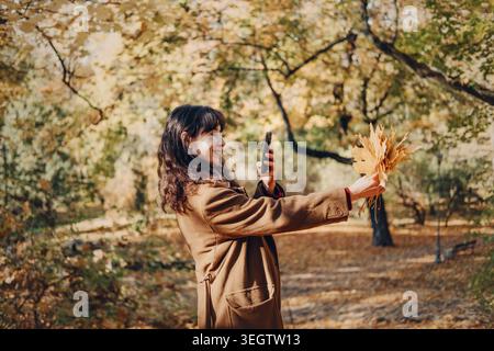 Person fotografiert gelbe Herbstblätter mit Smartphone im sonnigen Park. Fotografieren mit Smartphone, mobiles Fotografieren im Freien, weibliche Inhalte Stockfoto