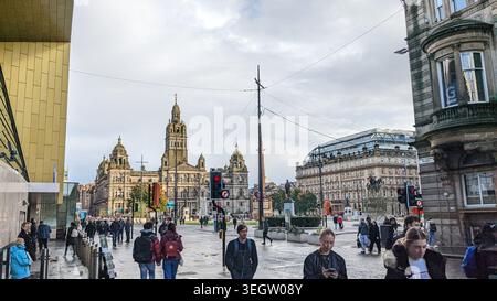 George Square im Stadtzentrum von Glasgow mit Fußgängern, die auf nassem Gehweg vor den historischen Glasgower City Chambers und dem umliegenden Architekten spazieren Stockfoto