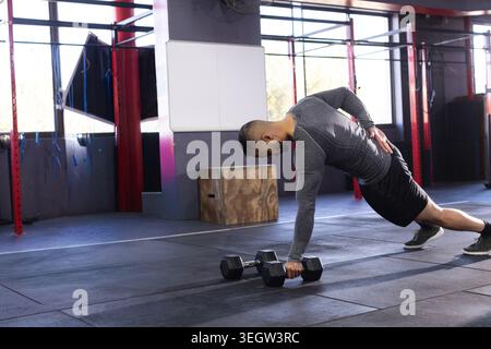 Indischer Mann Mitte der 30er Jahre, der eine Plank-Reihe mit Hexenhantel im Fitnessstudio mit grauem Kompressionsoberteil aufführt Stockfoto