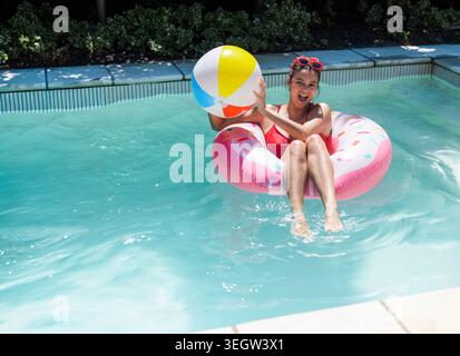 Frau, die in rosa Donut sitzt, schwimmt im türkisfarbenen Pool, hält einen bunten Ball, in rotem Badeanzug, Schatten Stockfoto