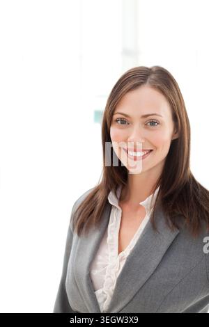 Geschäftsfrau lächelt und posiert im hellen Büro mit grauem Blazer und Rüschenbluse am Fenster Stockfoto