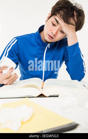 Ein junger Schüler sitzt an einem Schreibtisch, reibt sich die Stirn und hält ein Taschentuch, umgeben von Büchern und Papieren, sieht müde und gestresst aus. Stockfoto