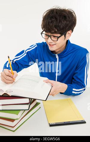 Ein junger Schüler sitzt an einem Schreibtisch, hält ein Buch und lächelt, während er am Rand schreibt. Stockfoto