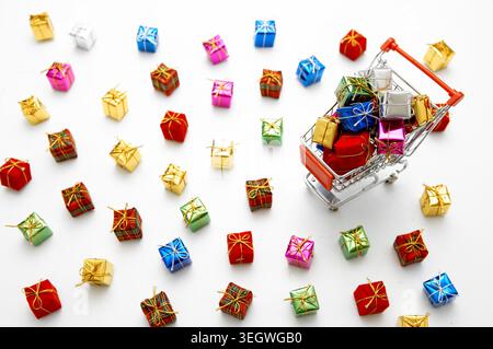 Ein Miniatur-Einkaufswagen mit kleinen Geschenkboxen ist umgeben von zahlreichen kleinen Geschenken auf weißem Hintergrund. Stockfoto