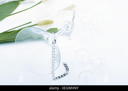 Eine silberne Kette und ein Glas Weinglas liegen auf einer weißen Oberfläche, umgeben von Wassertropfen und ein paar Blumen. Stockfoto