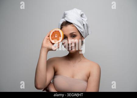 Junge Frau lächelt, trägt ein Handtuch Turban und hält eine Grapefruitscheibe nahe ihrem Gesicht, symbolisiert natürliche Hautpflege, Schönheit, Gesundheit und Wellness Stockfoto