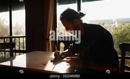 Mann, der digitale Kunst auf einem Tablet erstellt und von zu Hause aus mit natürlichem Licht arbeitet Stockfoto