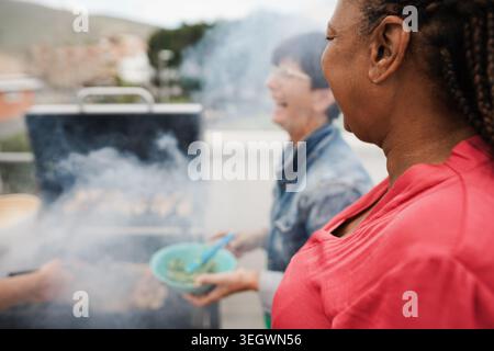 Multirassische Seniorenfreunde, die auf dem Dach zu Hause grillen - Essen, Sommer und Reife Menschen Lifestyle Konzept - Hauptfokus auf afrikanische Frau Ohr Stockfoto