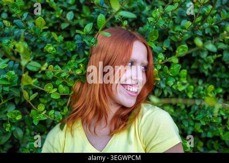Lächelnde rothaarige Frau mit fröhlichem Ausdruck und hellen Augenbrauen, die die Natur in einer üppigen grünen Umgebung genießt und ihre natürliche Schönheit zum Ausdruck bringt Stockfoto