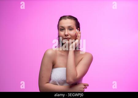 Eine junge Frau, die nach dem Duschen Gesichtscreme auf die Wange aufträgt, in ein weißes Handtuch mit nassem Haar gewickelt, um ihre tägliche Hautpflege gegen A zu üben Stockfoto