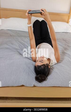 Indische Frau, die auf dem Bett liegt und die Tablette über dem Gesicht hält, während sie ein weißes Oberteil und ein Armband trägt Stockfoto