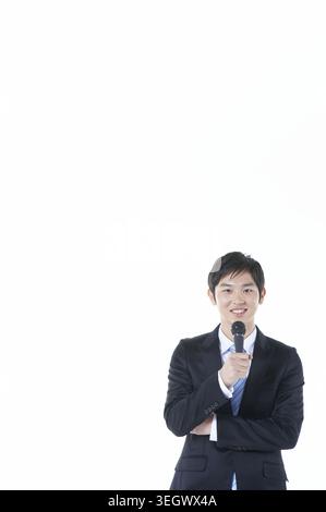 Ein junger Geschäftsmann in einem Anzug hält ein Mikrofon und spricht mit einer Menge in einer Konferenz oder öffentlichen Sprechstunde. Stockfoto