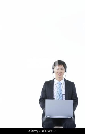 Ein Geschäftsmann sitzt mit einem Laptop auf dem Schoß und trägt Kopfhörer in einer neutralen Büroumgebung. Stockfoto