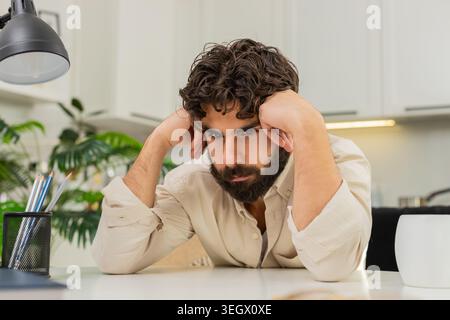 Der indische Mann sitzt zu Hause still und fühlt Burnout keine Inspiration geringe Motivation müde geistig schwer. Ein arabischer Typ, der am Tisch sitzt, schaut nach unten langsam atmende Hände auf die Wangen empfindet Stress Traurigkeit geistige Ermüdung. Stockfoto