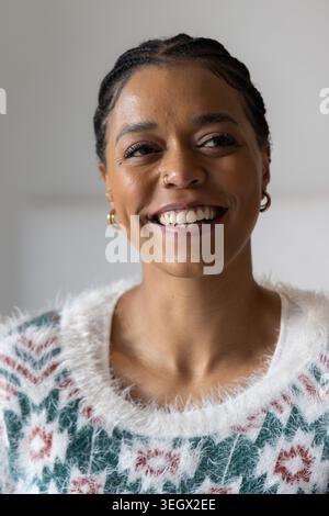 Erwachsene afroamerikanische Frau lächelt und blickt zu Hause in flauschigem Pullover geflochten goldenen Creolen Nasenring Stockfoto