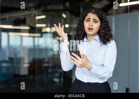 Indische Geschäftsfrau steht in einem Büro, hält ein Smartphone in der Hand, sieht frustriert und verwirrt aus, hat Probleme, schlechte Nachrichten zu verstehen oder einen technischen Fehler auf ihrem Gerät Stockfoto