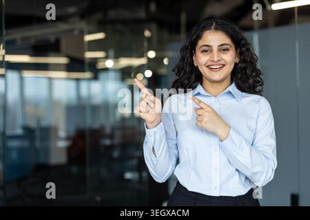 Lächelnde Geschäftsfrau in einem hellblauen Hemd, die beide Zeigefinger nach links zeigt, etwas mit fröhlichem Ausdruck in einer modernen Büroumgebung zeigt, präsentiert oder zeigt Stockfoto