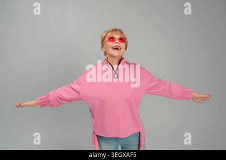 Eine Erwachsene, emotionale, schelmische, glückliche Frau in rosa Kleidung und Brille mit erhobenen Händen Stockfoto