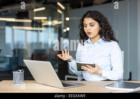 Junge indische Geschäftsfrau, die einen Laptop für einen Online-Videoanruf in ihrem Büro benutzt und sich über potenzielle technische Probleme gestresst und verwirrt fühlt, während sie ein Notebook hält Stockfoto