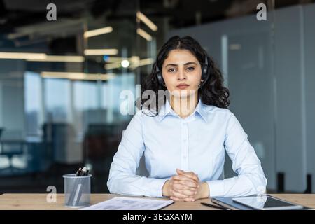 Junge Geschäftsfrau mit dunklen lockigen Haaren, die ein Headset und ein hellblaues Hemd trägt, mit gefalteten Händen an einem Schreibtisch sitzt und in einem modernen Callcenter-Büro als Contact Center-Operator arbeitet Stockfoto