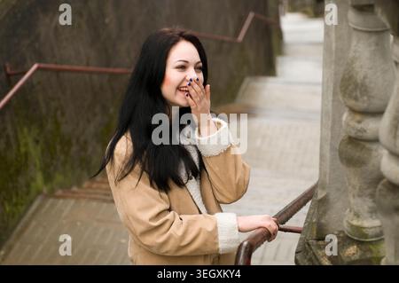 Junge Frau lacht und bedeckt ihren Mund, während sie an einem Geländer auf einer Außentreppe aus Stein steht Stockfoto