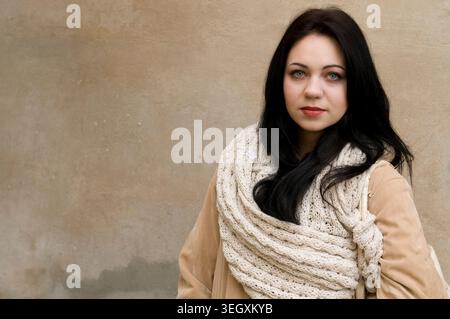 Junge Frau in einem beigen Mantel und einem dicken gestrickten Schal, die vor einer schlichten neutralen Wand posiert Stockfoto