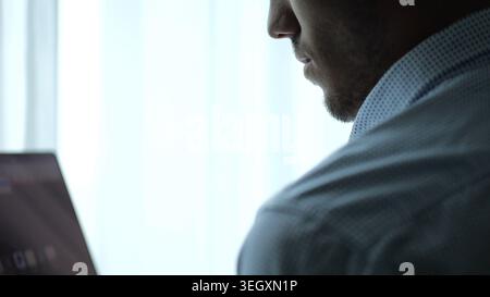 Nahporträt eines jungen Unternehmers, der in der Nähe eines großen Fensters in einem modernen Büro steht und an einem Notebook arbeitet. Hübscher Geschäftsmann im Hemd Stockfoto