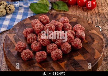 Eine Nahaufnahme roher, ungekochter Fleischbällchen auf einem dunklen Holzschneidebrett, umgeben von frischen Basilikumblättern, roten Kirschtomaten, Wachteleiern Stockfoto
