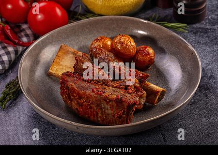 Appetitliche gegrillte Rinderrippchen glasiert mit Senfsauce serviert mit gebratenen Kartoffeln in einer rustikalen grauen Schüssel. Herzhaftes Gourmet-Essen auf dunklem Backgrou Stockfoto
