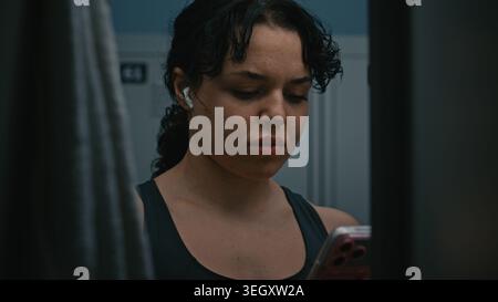 Fokussierte junge Athletin zieht kabellose Kopfhörer an und überprüft das Smartphone vor dem Workout im Schließfach. Moderner Fitness Lifestyle, Motivation, Musik und Vorbereitung auf intensive Trainingseinheiten. Stockfoto
