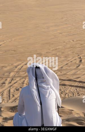 Eine Rückansicht einer Person, die einen traditionellen weißen Thobe und Gghutra trägt und auf einem Sanddünenhang bei Khor Al Adaid in Al Wakrah, Katar, sitzt. Stockfoto