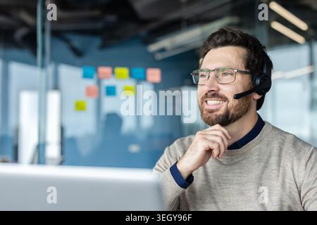 Glücklicher bärtiger Mann mit Brille und Headset, der am Laptop arbeitet, Unterstützung und Unterstützung bietet und ein positives Kundenerlebnis in einer modernen Büroumgebung verkörpert Stockfoto