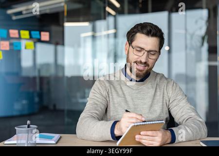 Junger, lächelnder Geschäftsmann mit Brille, der an einem modernen Schreibtisch in ein Spiralnotizbuch schreibt und sich auf Brainstorming, die Planung von Aufgaben und die Organisation von Ideen für ein Projekt oder eine Besprechung konzentriert Stockfoto