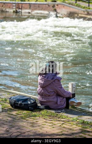 Junge asiatische Frau, die allein am Ufer des Flusses Cherwell in Reading, Berkshire, England, Großbritannien sitzt Stockfoto