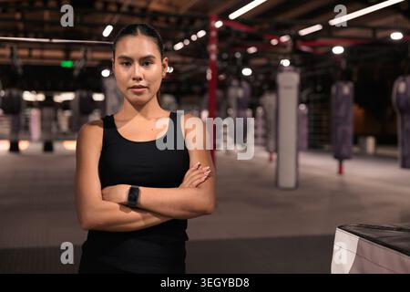 Ein horizontaler mittlerer Schuss einer entschlossenen jungen asiatischen Athletin, die mit überkreuzten Armen in einem modernen Boxstudio posiert. Sie trägt einen schwarzen Panzer Stockfoto