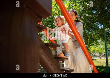 Mutter und Sohn des Kleinkindes teilen einen freudigen Moment, während sie ihn an einem hellen sonnigen Tag in einem hölzernen Spielhaus in einem grünen Park führt und Wärme und Vertrauen einfängt Stockfoto