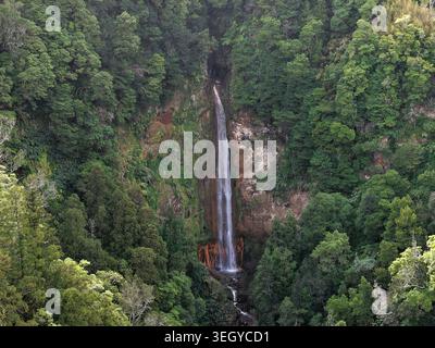 Blick aus der Vogelperspektive auf den Wasserfall Cascata da Ribeira Quente, der eine steile Klippe hinabstürzt, umgeben von üppigen grünen Wäldern in Ribeira Quente, Azoren, Portugal. Stockfoto