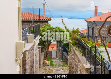 Enge gepflasterte Straße, die sich zwischen traditionellen Häusern in Molyvos, Lesbos, Griechenland, schlängelt und mit Ziegeldächern, üppigem Grün, und einen malerischen Meerblick Stockfoto