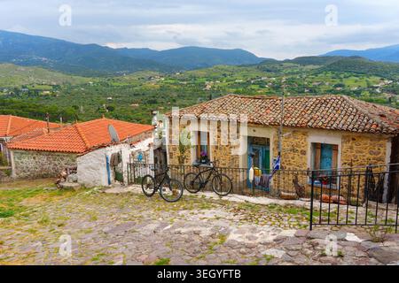Traditionelles Steinhaus mit Terrakotta-Ziegeldach und Fahrrädern, die draußen auf einer kopfsteingepflasterten Straße im Dorf Molyvos, Lesbos, Griechenland, mit Blick auf den Ort geparkt werden Stockfoto